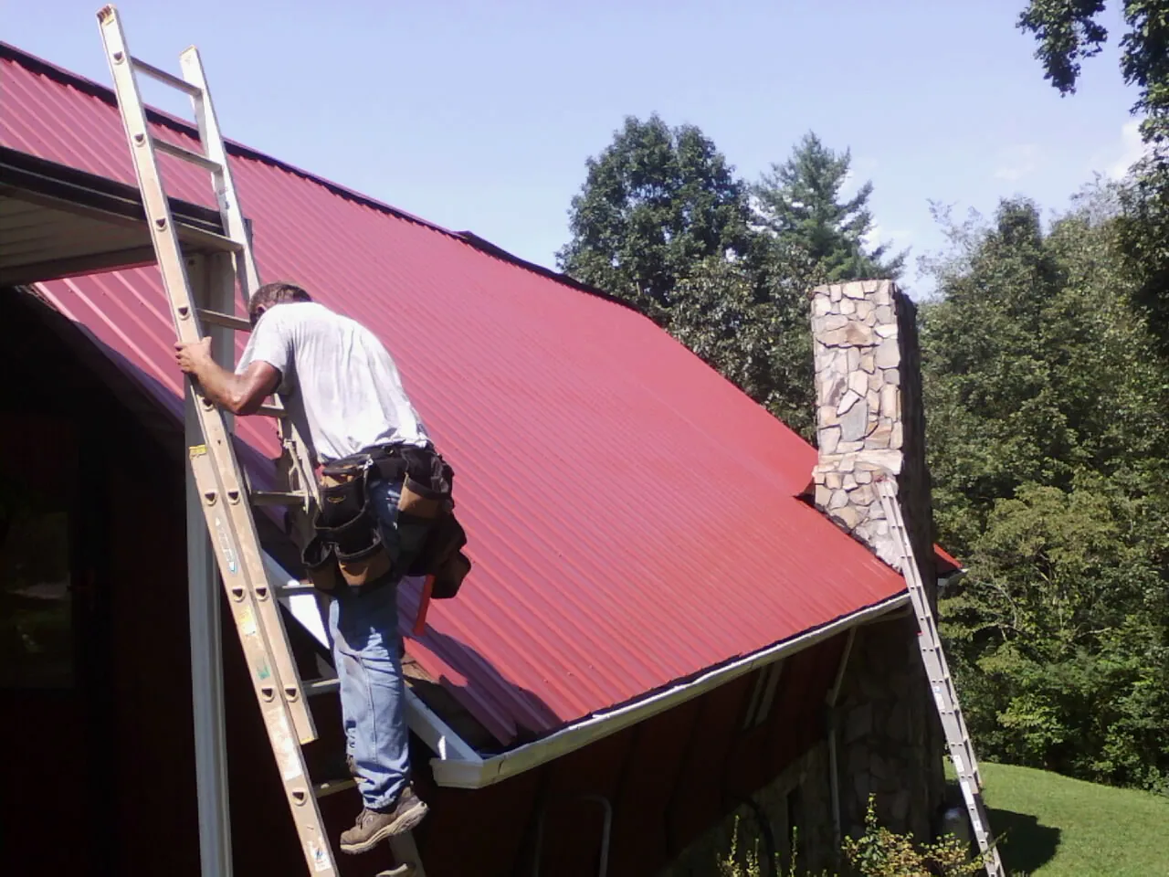 Roofer on a ladder beside a red metal roof and stone chimney — Soffit & Fascia Repair by Roofing Brothers in Tabor City